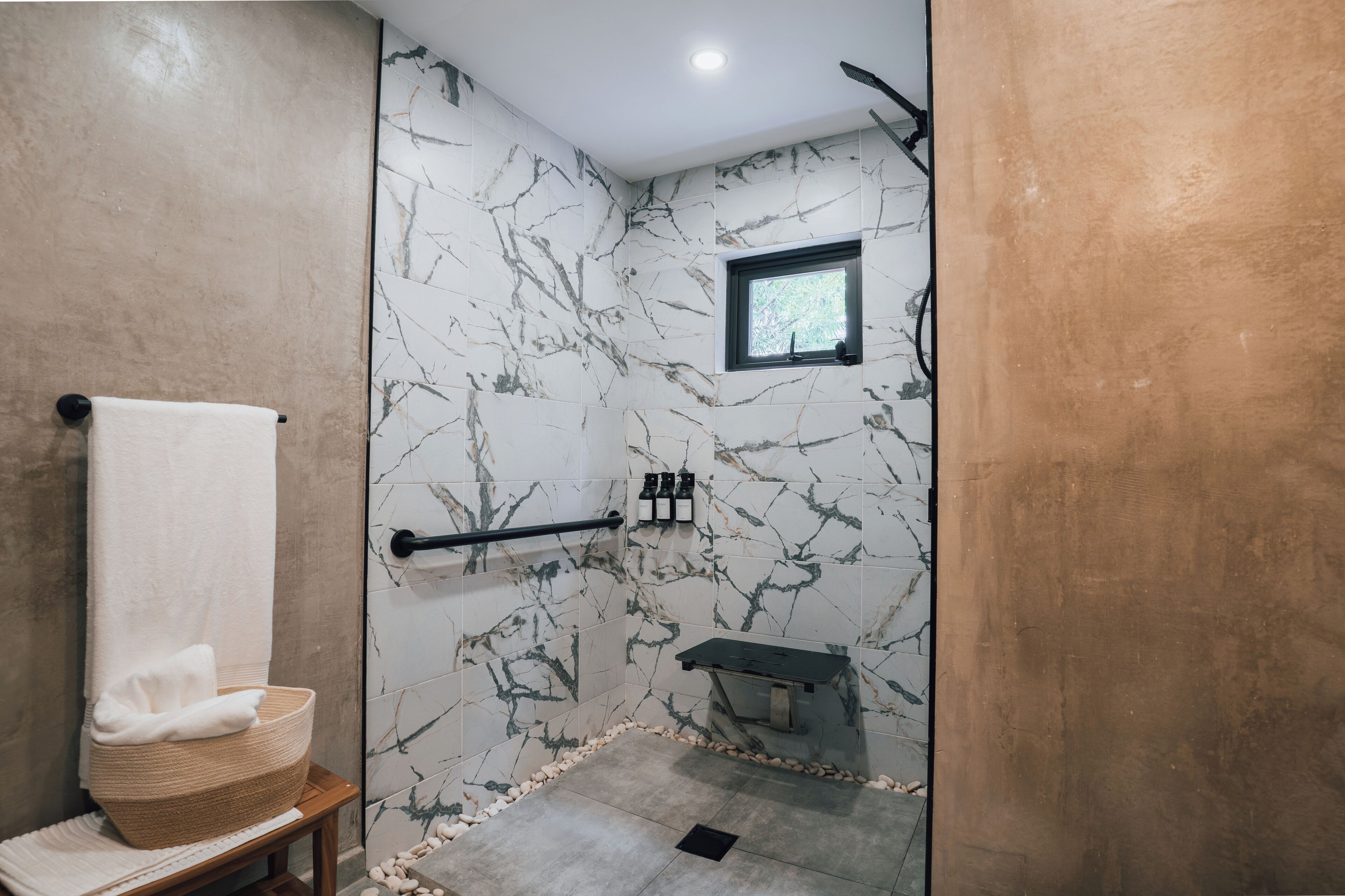 A modern bathroom with a marble wall, a small window, a towel rack, and a bench.