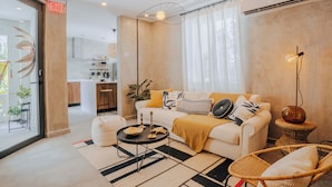 A modern living room with a white sofa, a round coffee table, and a wicker chair. There is a kitchen in the background with wooden cabinets and a bar stool. - Flor De India (San Juan)