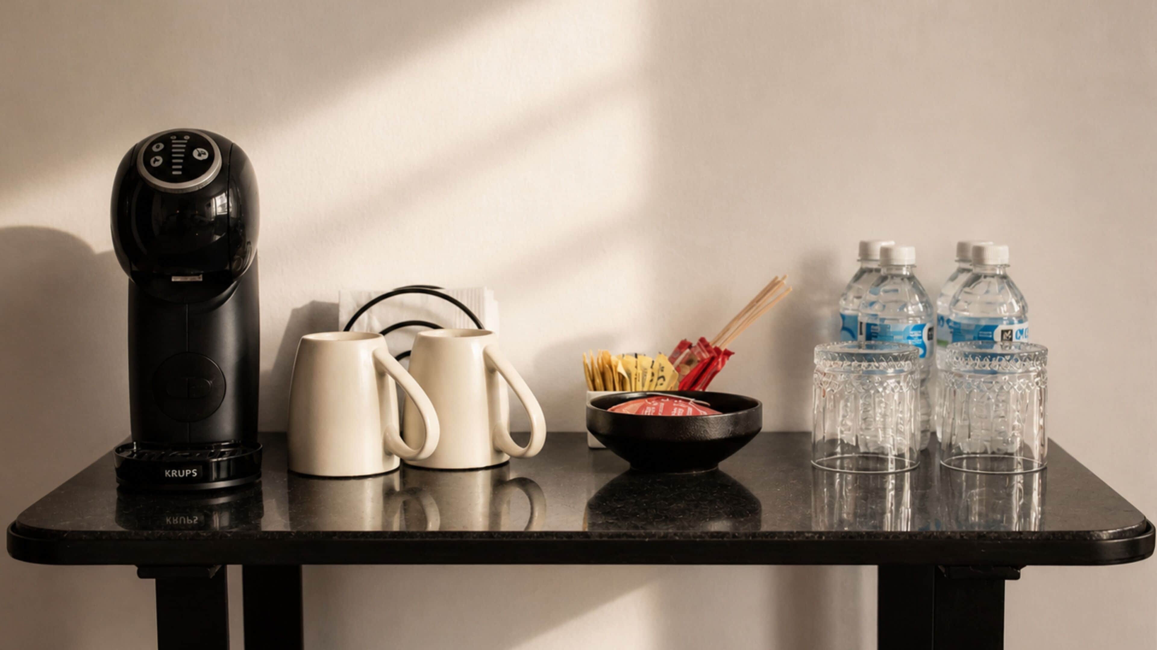 Una mesa con una cafetera, dos tazas, un bol de aperitivos y dos botellas de agua.
