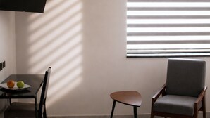 A modern living room with a flat-screen TV mounted on the wall, a dining table with chairs, a small stool, and a window with blinds.