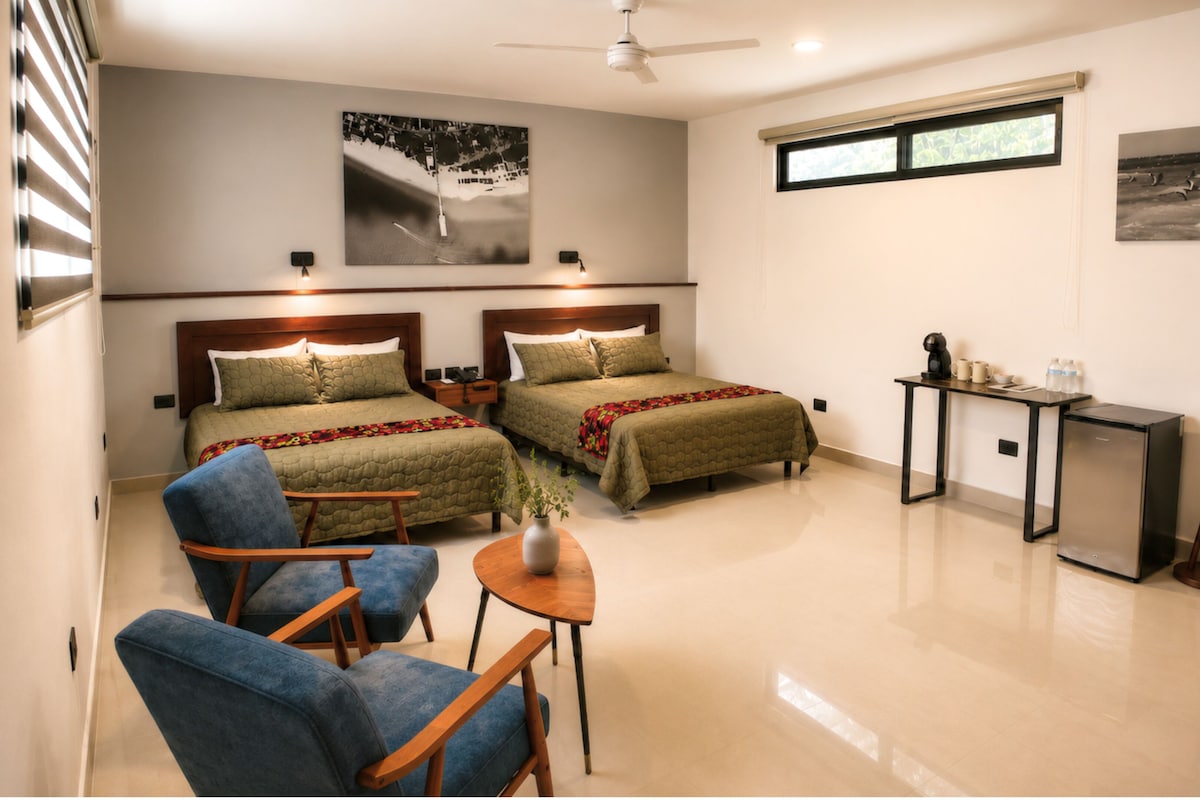 A hotel room with two beds, a desk, a chair, a small table, and a plant.