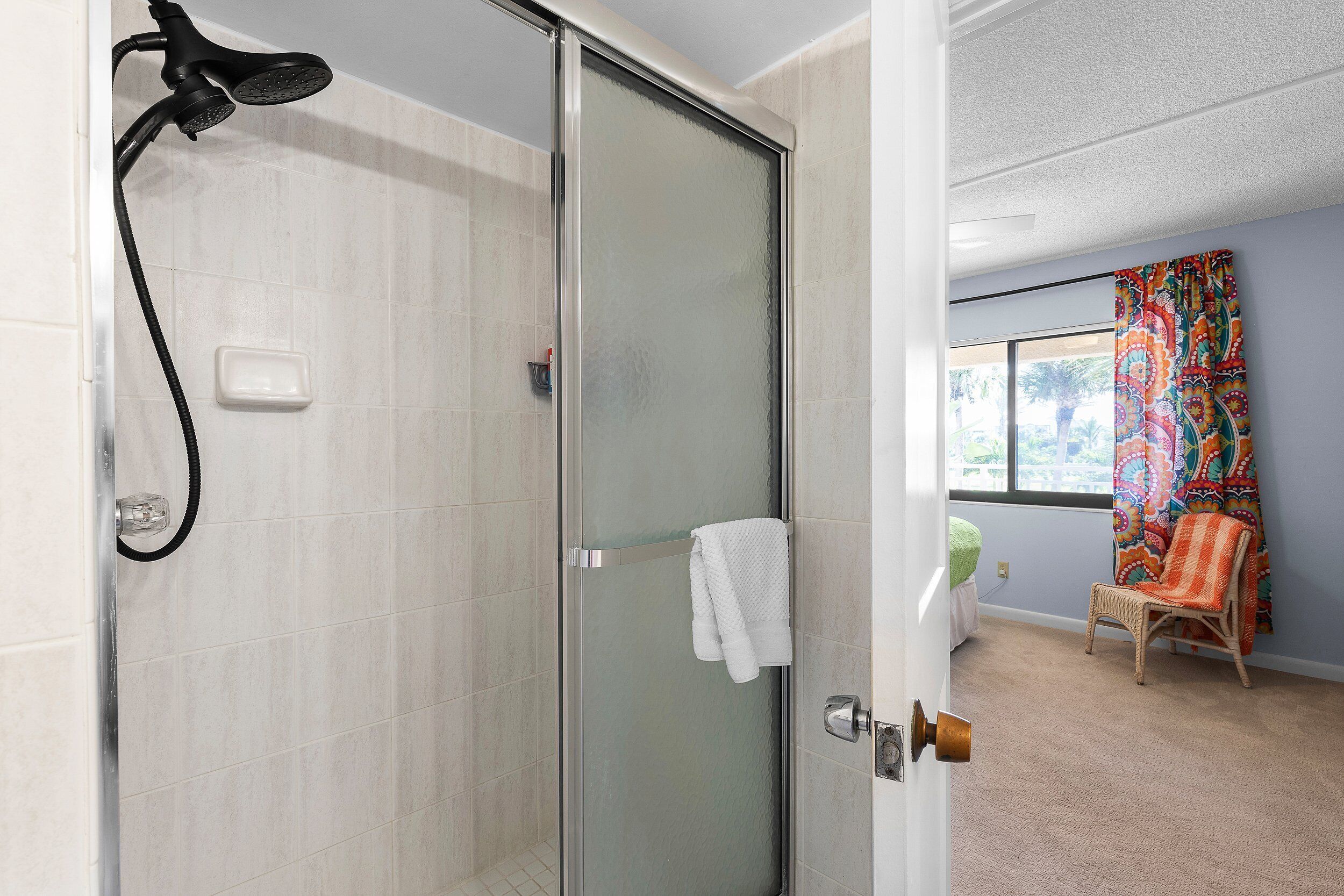 A modern bathroom with a glass-enclosed shower, a white towel hanging on a rack, and a wicker chair in a room with a large window and colorful curtains.