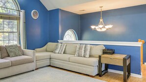 A room with a beige sectional sofa, a small side table, a large window with a view of trees, and a blue accent wall. - Hike, Ski & Explore: Otisville Retreat w/ Yard! (Otisville)