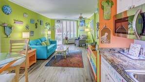 A bright living room with a teal sofa, a glass dining table, and a kitchen with a 'Welcome to Our Space' sign. - Oceanfront Myrtle Beach Condo: Resort Perks! (Myrtle Beach)
