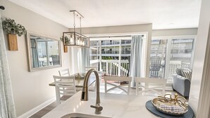 A modern kitchen with a stainless steel sink, white cabinetry, and a dining area with a white table and chairs. - Sandpiper Cove Unit 1027  1 Bedroom With FREE Beach Service (Destin)