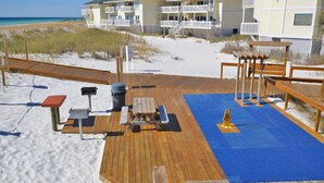 Eine Holzterrasse mit Picknicktischen und Grill, mit Blick auf einen Strand und ein Gebäude mit Balkonen.