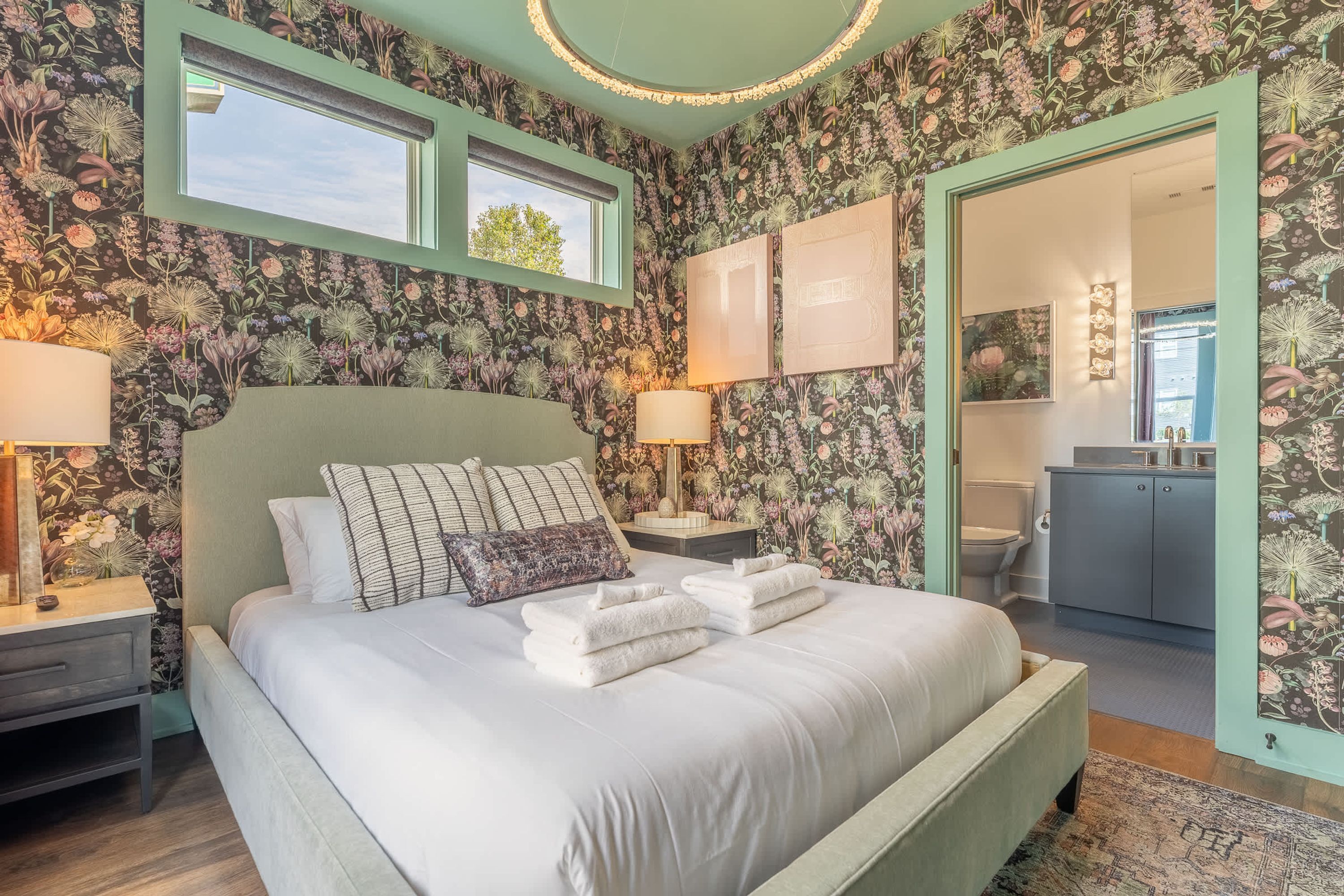 A bedroom with a floral wallpaper, a bed with white linens, two bedside lamps, and a view of the bathroom through an open door.