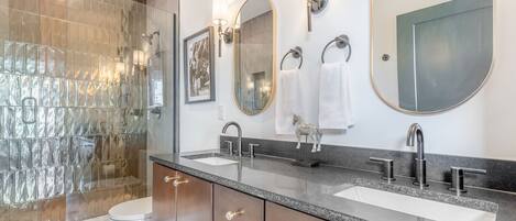 A modern bathroom with a glass shower enclosure, two sinks, a large mirror, and wall-mounted lighting.