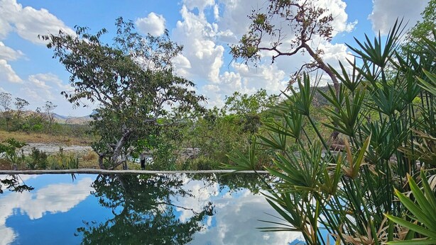 A serene outdoor scene with a clear sky, a calm body of water reflecting the sky, and a small dog resting on the ground near some plants. - Camping Solar da Colina (Colinas do Sul)