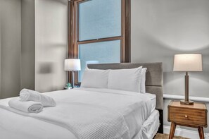 A neatly made bed with white linens and a rolled towel, a bedside lamp, and a nightstand with a drawer.