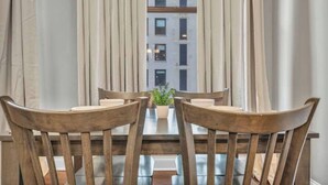 Una mesa de comedor con cuatro sillas de madera, una planta en maceta y vistas de un edificio a través de cortinas translúcidas.