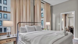 Habitación de hotel con una cama grande, una mesita de noche con lámpara, un espejo y vistas a un edificio.