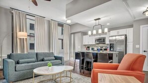 Una sala de estar moderna con un sofá gris, un sillón naranja, una mesa de centro redonda y una cocina al fondo.