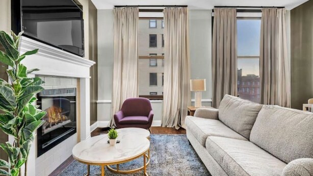 A modern living room with a fireplace, a sofa, a round coffee table, and a view of the city through large windows. - Luxury Loft • Heart of Downtown• FREE Parking/Gym (Cleveland)