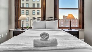 A hotel room with a large bed, two bedside lamps, a window with a view of buildings, and a towel on the bed. - Luxury Loft • Heart of Downtown• FREE Parking/Gym (Cleveland)