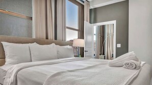 Ein Hotelzimmer mit einem großen Bett, einem Nachttisch, einer Lampe und Blick auf die Stadt durch das Fenster.