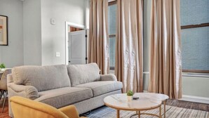 Un salón con un sofá gris, una mesa de centro redonda y un sillón amarillo. Se ve una ventana con cortinas y una puerta al fondo.