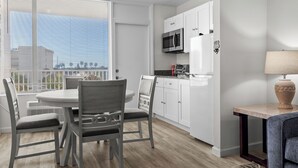 A modern kitchen with white cabinets, a microwave, and a refrigerator. There is a dining table with chairs and a window with blinds.