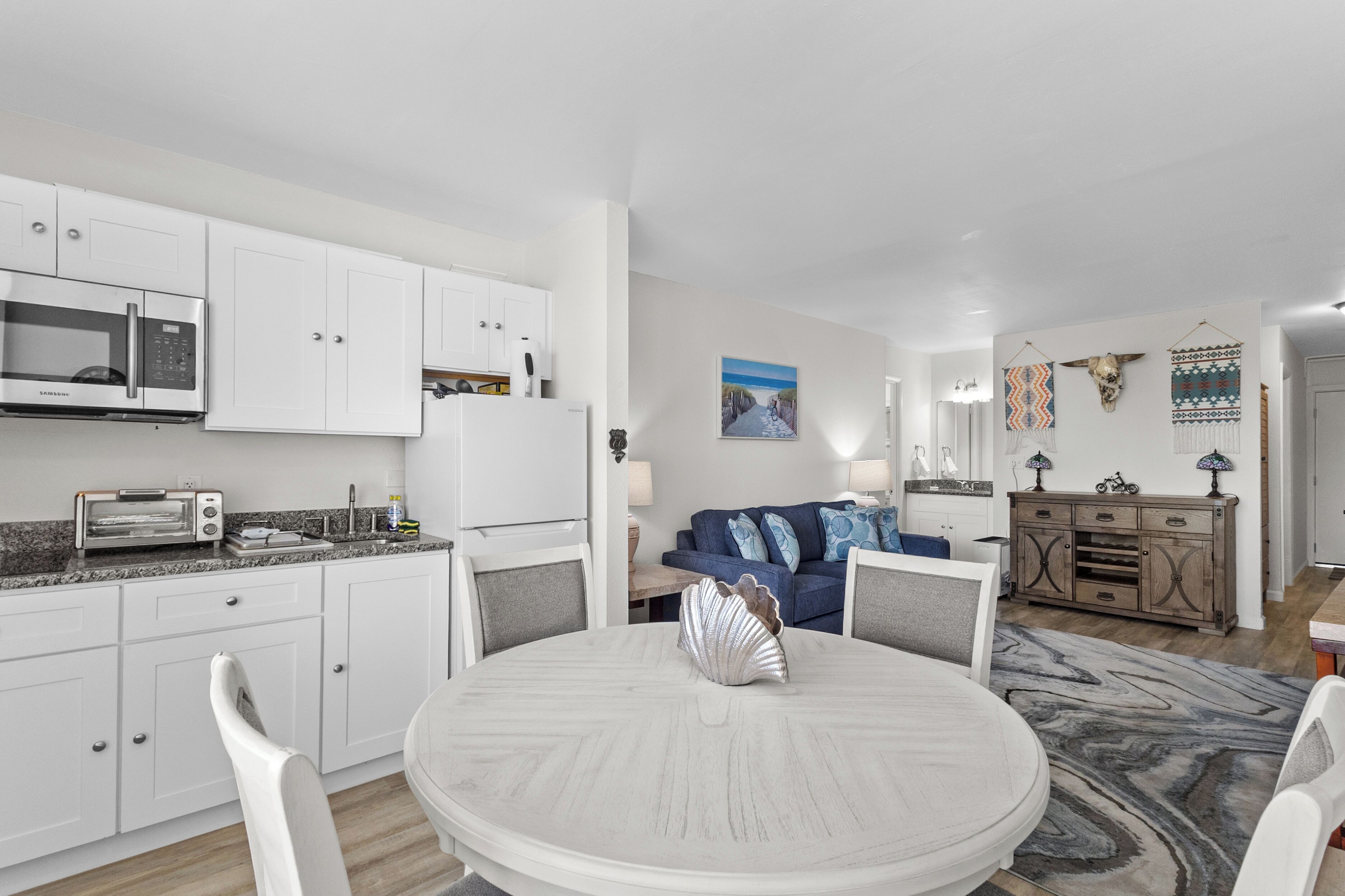 Una cocina moderna con armarios blancos, microondas y tostadora. Hay una mesa de comedor con sillas y un centro de mesa decorativo con una concha.