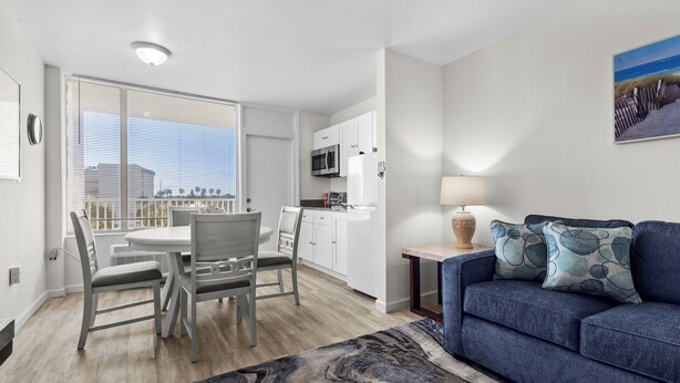 A modern living room with a blue sofa, a dining table with chairs, a kitchen area with white cabinets, and a large window with blinds.
