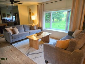 A living room with a sofa, armchair, coffee table, and a window with a view of greenery.