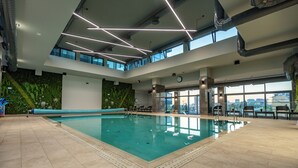 Ein modernes Hallenbad mit freiem Blick auf die Stadt durch große Fenster, einer begrünten Wand mit Pflanzen und einer Sitzecke mit Stühlen und einer Bank.