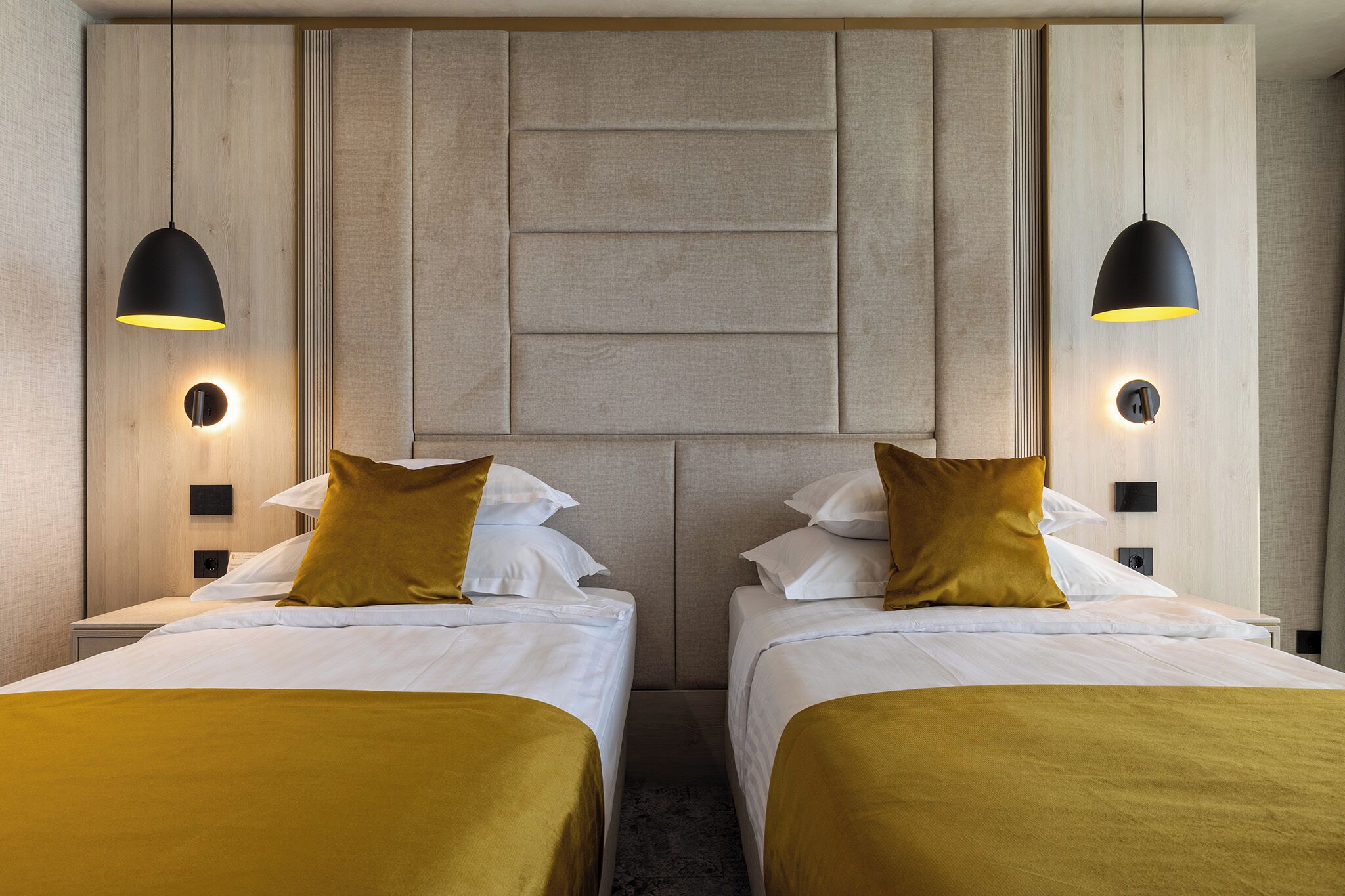 A hotel room with two beds, each with a headboard, a central wall panel, and two pendant lights.
