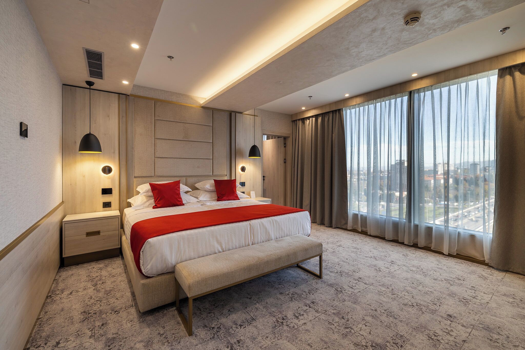 A modern hotel room with a large bed, a bench, a nightstand, and a view of the city through large windows.