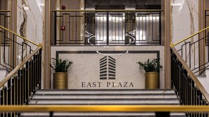 Eine Marmortreppe führt zu einem Aufzug. An diesem befindet sich der Schriftzug "EAST PLAZA" und je einer getopfte Pflanze auf jeder Seite.