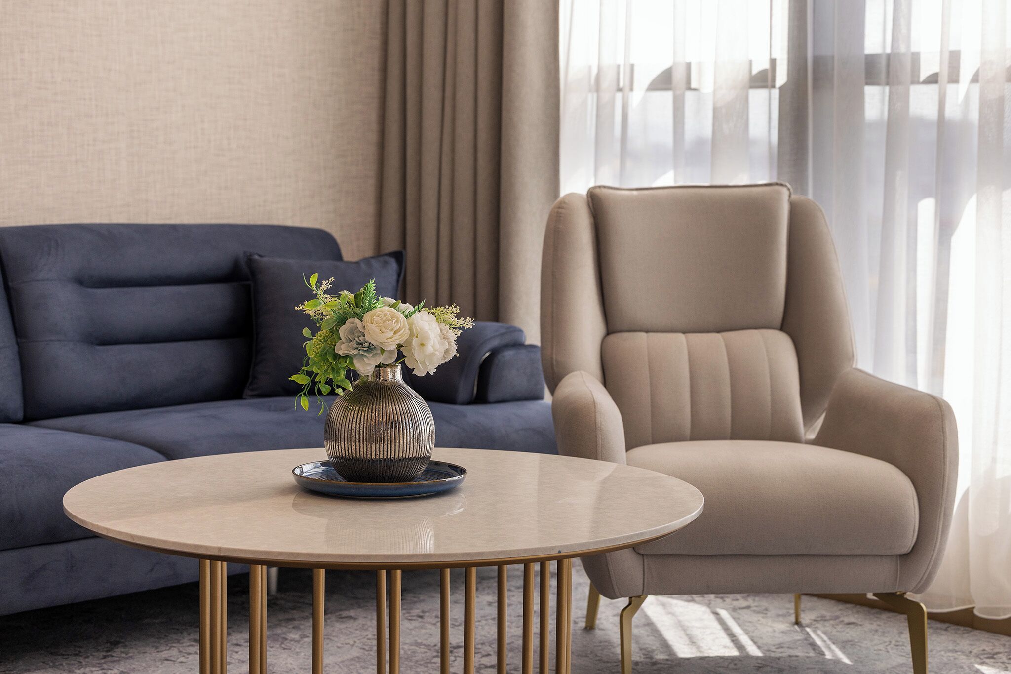 Una sala de estar moderna con un sofá azul oscuro, un sillón beige y una mesa redonda de centro con un jarrón de flores.