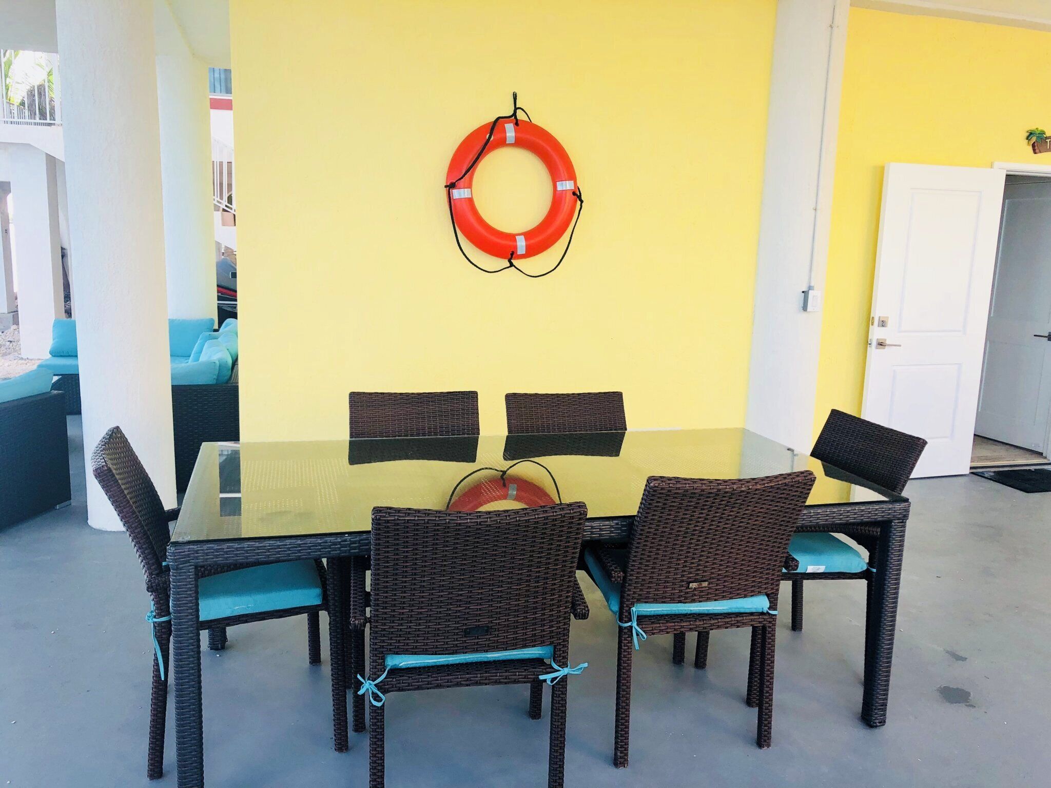 Un coin repas avec une table en verre entourée de chaises en osier, un mur jaune et un gilet de sauvetage accroché au mur.