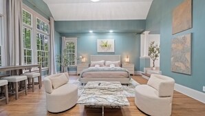 A bedroom with a bed, two armchairs, a coffee table, a desk with chairs, and a large window.