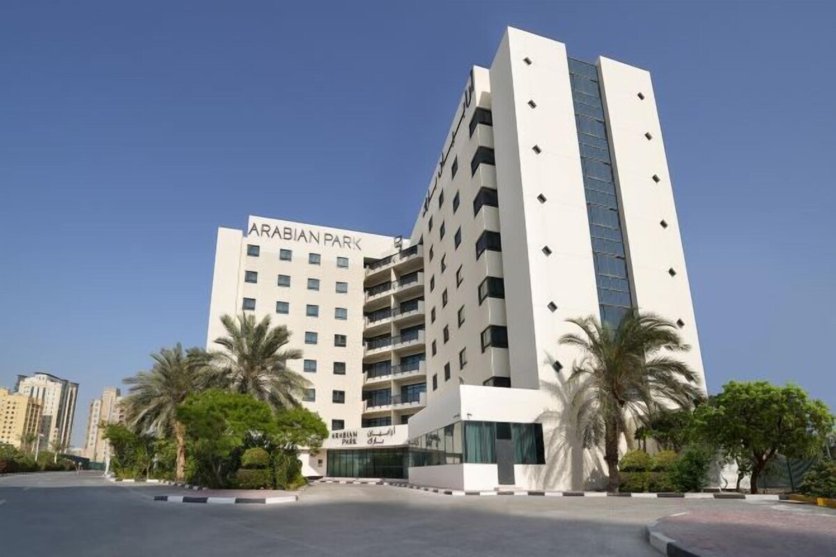 Un edifici modern de diversos pisos amb el senyal 'ARABIAN PARK', envoltat de palmeres i un cel serè.