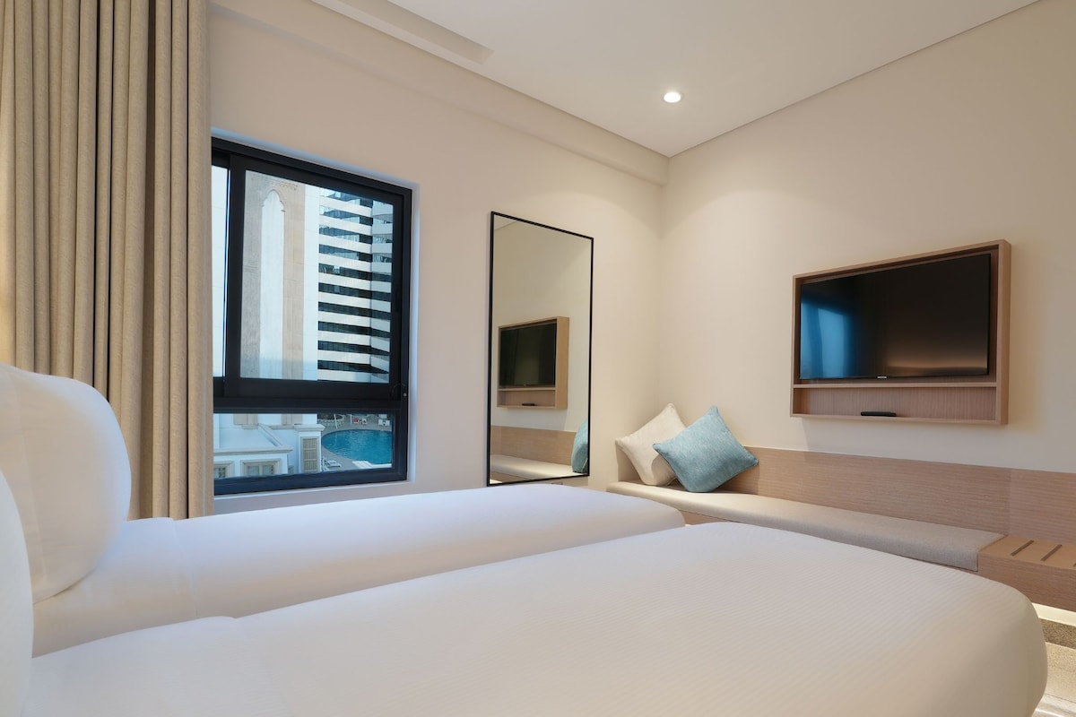 Une chambre d’hôtel avec un grand lit, un téléviseur fixé au mur et une fenêtre donnant sur un immeuble et une piscine.
