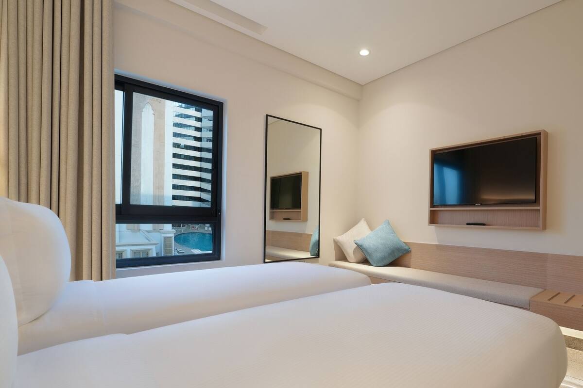 Une chambre d’hôtel avec un grand lit, un téléviseur fixé au mur et une fenêtre donnant sur un immeuble et une piscine.