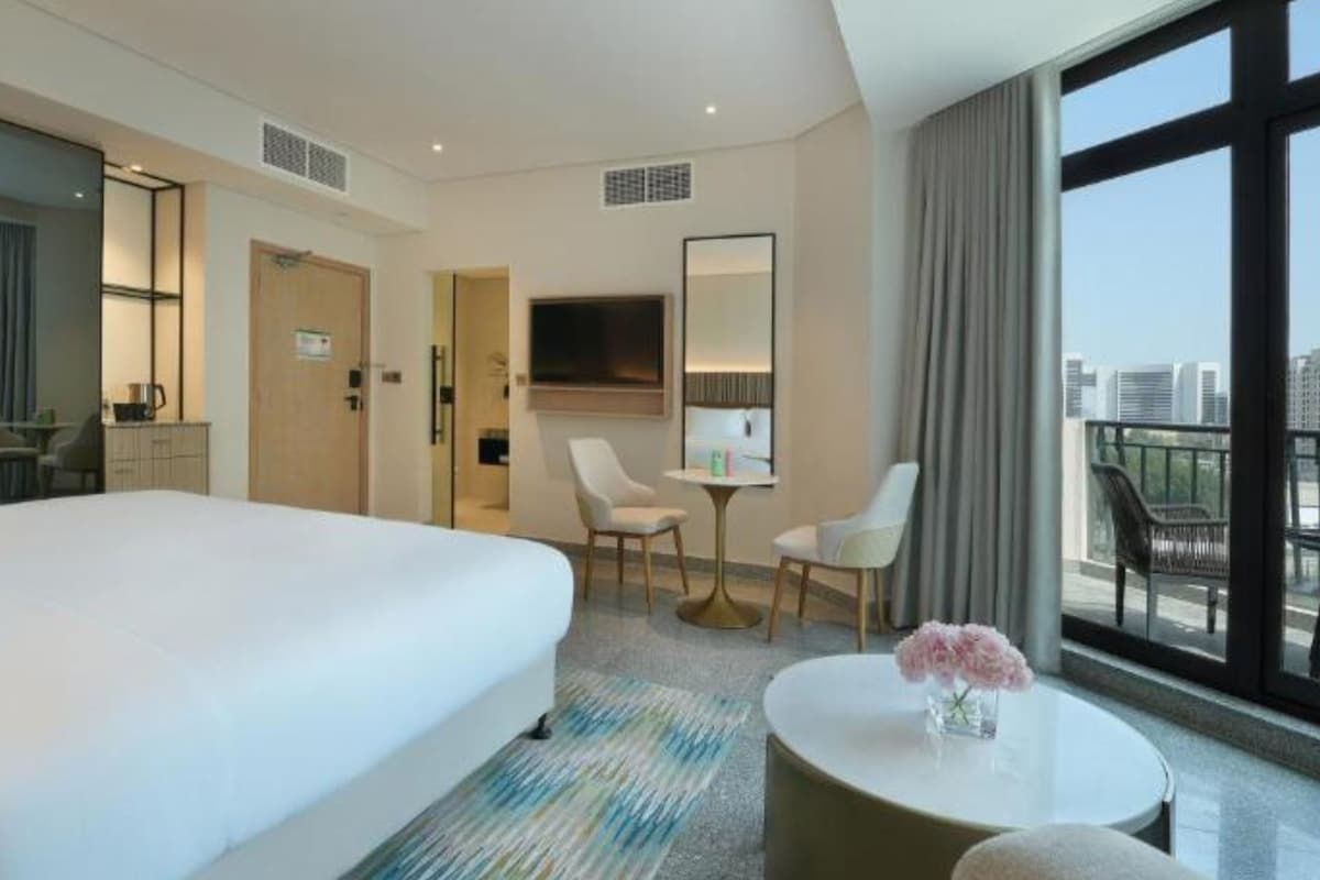 Une chambre d’hôtel avec un grand lit, un bureau avec une chaise, une petite table ronde avec une chaise, une télévision et une grande fenêtre donnant sur la ville.