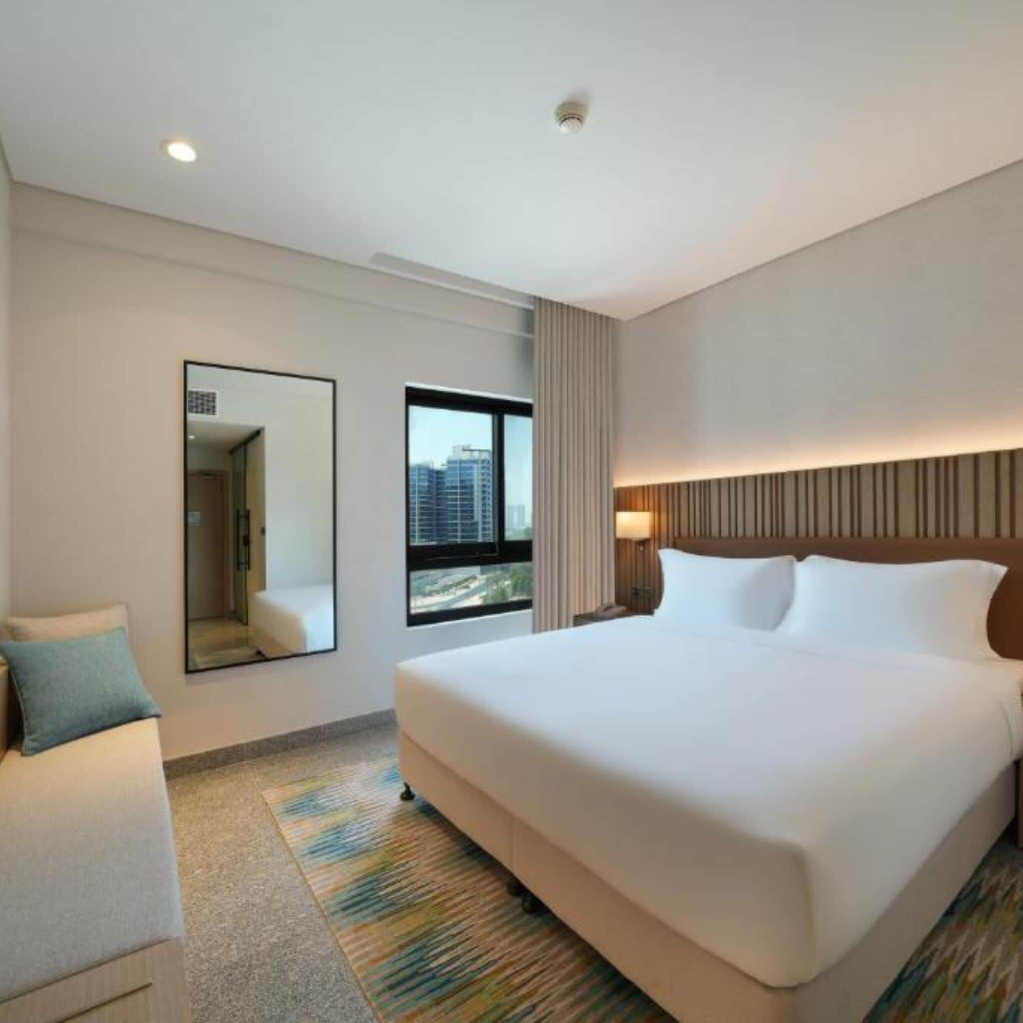 A hotel room with a large bed, a mirror, a window with a city view, and a ceiling light.