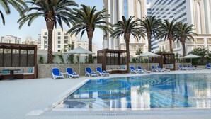 Un área de piscina con sillones de descanso, palmeras y edificios modernos al fondo.
