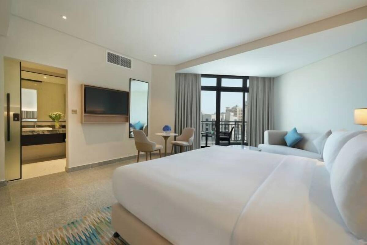 Une chambre d’hôtel dotée d’un grand lit, d’un téléviseur fixé au mur, d’un bureau avec des chaises et offrant une vue sur la ville grâce à de grandes fenêtres.