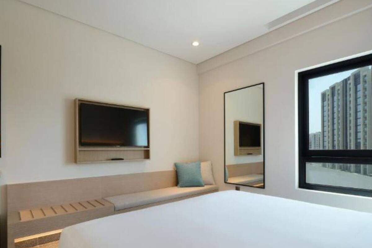 Une chambre d’hôtel moderne dotée d’un grand lit, d’un téléviseur à écran plat fixé au mur, d’une grande fenêtre offrant une vue sur la ville et d’une banquette intégrée avec espace de rangement.