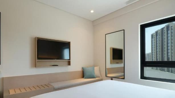 Una habitación de hotel moderna con una cama grande, un televisor de pantalla plana en la pared, un ventanal con vista a la ciudad y un banco empotrado con espacio de almacenamiento.