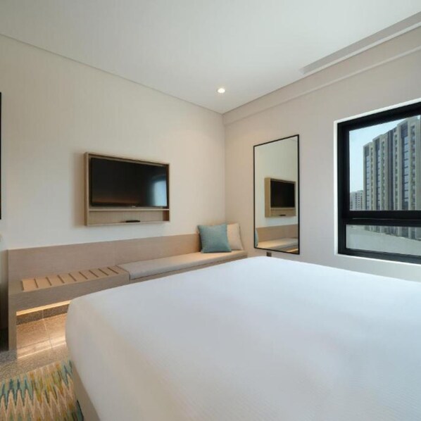 Une chambre d’hôtel moderne dotée d’un grand lit, d’un téléviseur à écran plat fixé au mur, d’une grande fenêtre offrant une vue sur la ville et d’une banquette intégrée avec espace de rangement.