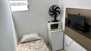 A small, narrow room with a bed, a microwave, a fan, and a TV mounted on the wall. - Quarto individual em São Paulo ZN (São Paulo)