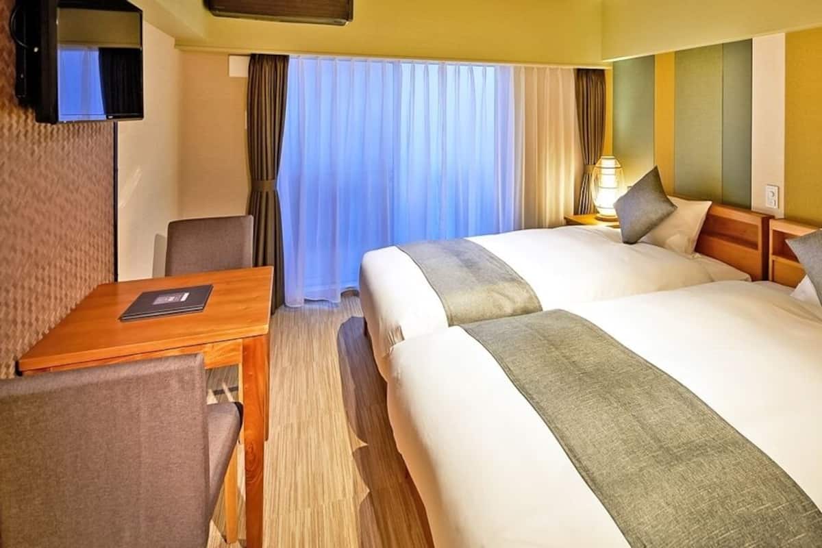 Ein Hotelzimmer mit zwei Betten, einem Schreibtisch, einem Fernseher und einem Fenster mit Vorhängen.