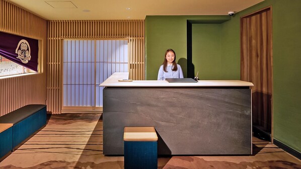 A reception desk with a person seated behind it, a wooden bench, and a large window with vertical blinds. - Stay Sakura Tokyo Asakusa Yokozuna Hotel (Tokyo)