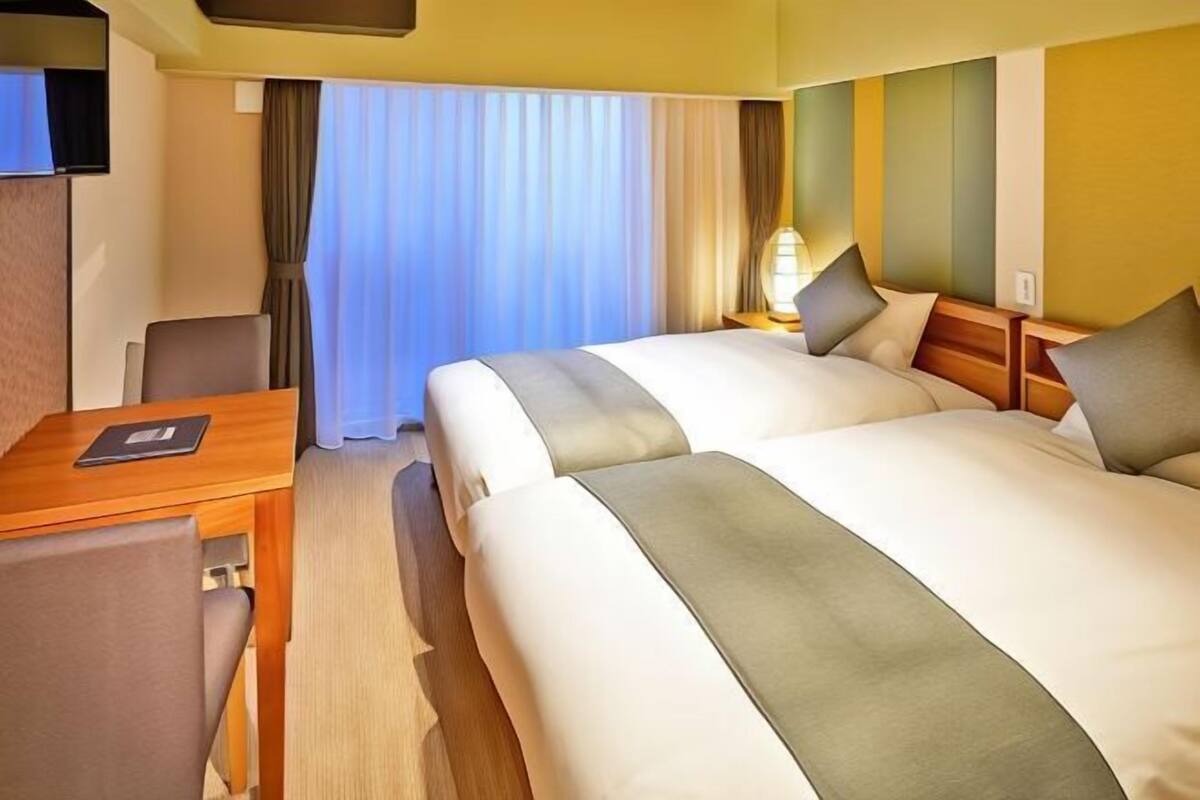 Ein Hotelzimmer mit zwei Betten, einem Schreibtisch, einem Stuhl, einem Fernseher und einem Fenster mit Vorhängen.