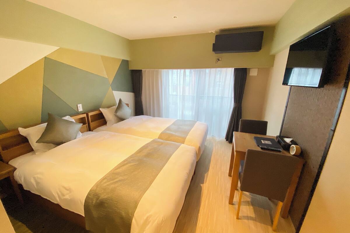 Ein Hotelzimmer mit zwei Betten, einem Schreibtisch, einem Sessel, einem Fernseher und einem Fenster mit Vorhängen.