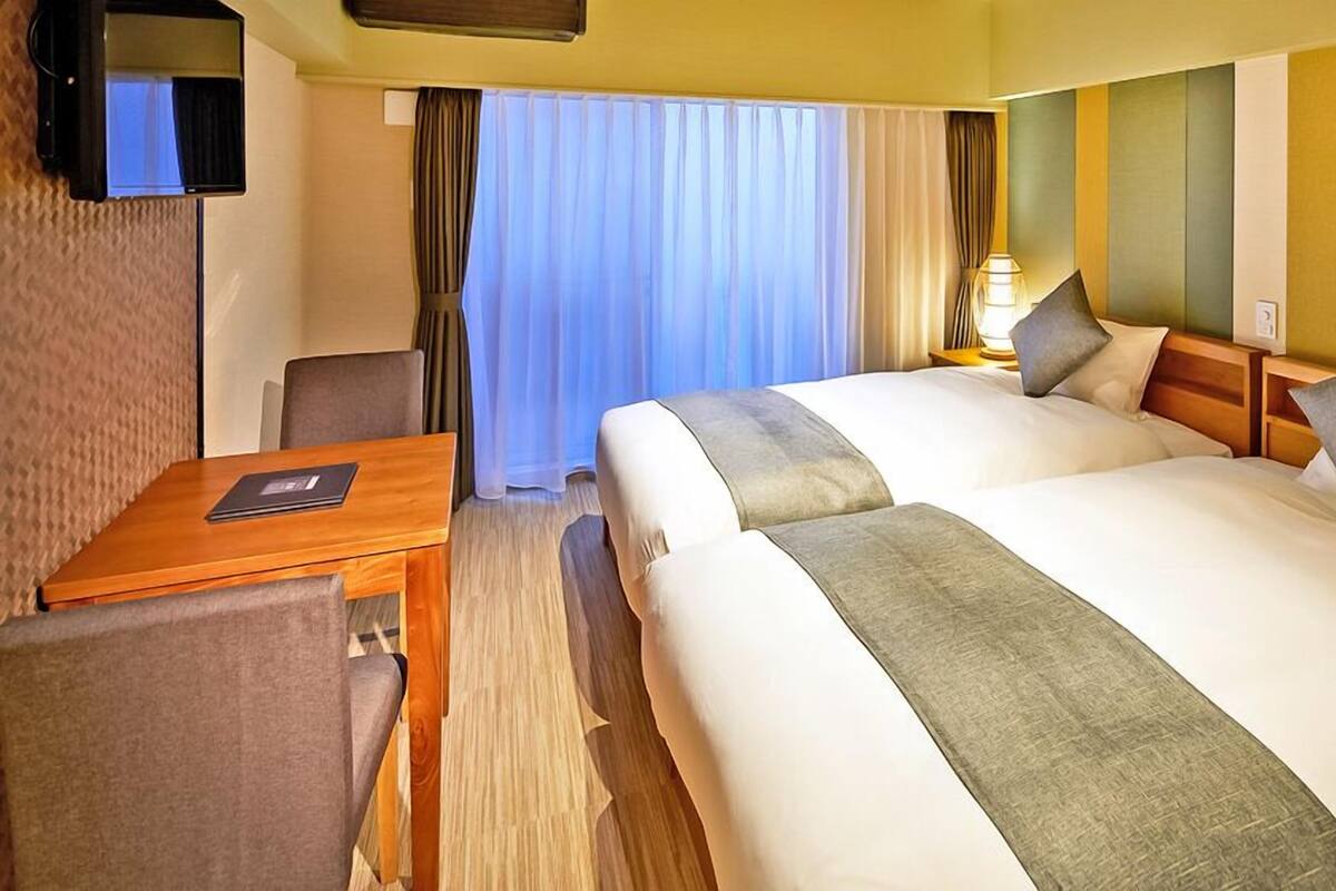 Ein Hotelzimmer mit zwei Betten, einem Schreibtisch, einem Stuhl, einem Fernseher und einem Fenster mit Vorhängen.