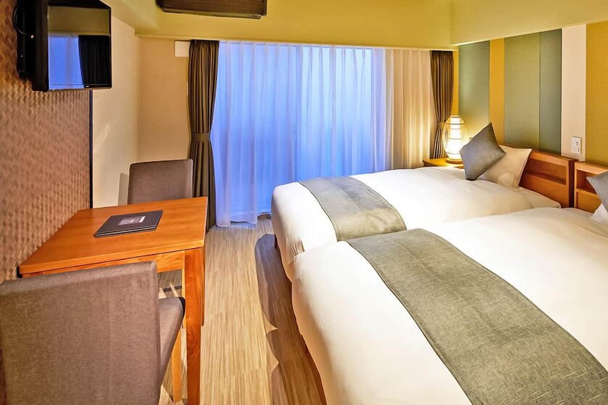 A hotel room with two beds, a desk, a chair, a TV, and a window with curtains.