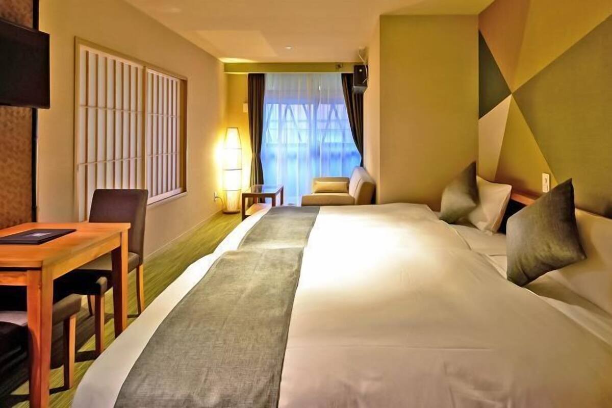 Ein Hotelzimmer mit Bett, Schreibtisch, Stuhl, Lampe und einem fensterbepflanzten Fenster.
